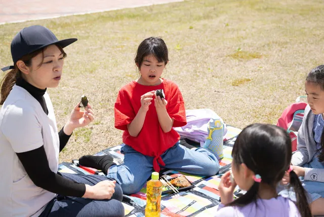 遠足でお弁当を食べる先生と小学生
